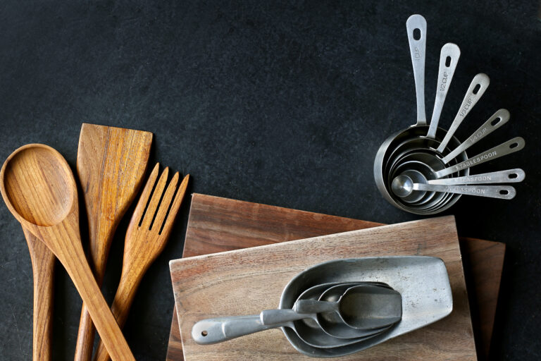 A collection of vintage baking and cooking supplies, wood cutting boards and spoons, and antique silver measuring cups frame a black slate chalkboard background.